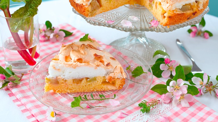 Ein Stück Rhabarber-Baiser-Kuchen auf einem Glas-Teller vor einer Kuchenplatte mit weiteren Kuchenstücken, dekoriert mit Blüten.