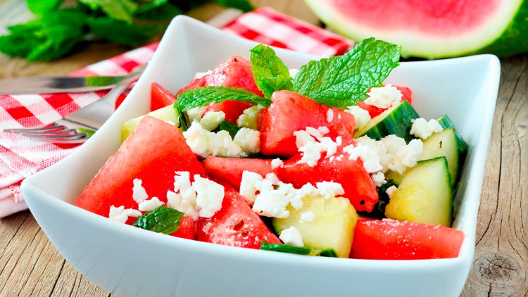 Würfeln aus Wassermelone, Gurke, Feta und Minzblättern in einer weißen eckigen Schale auf Holztisch mit rotem Stofftuch.