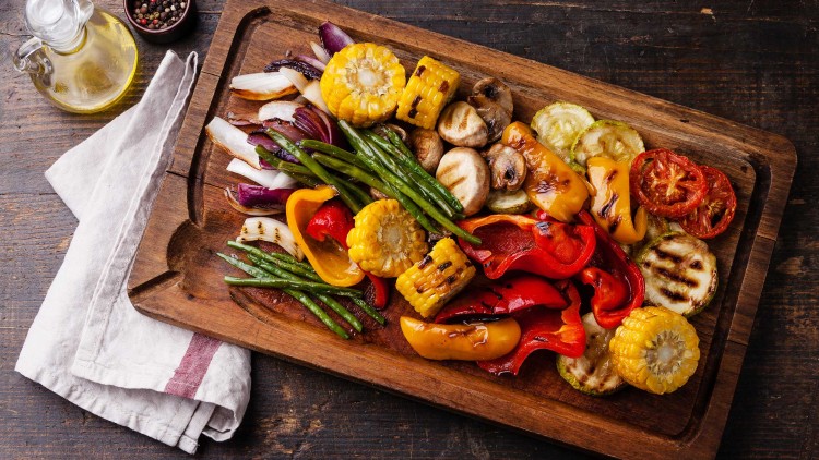 Gegrilltes Gemüse wie Mais, Paprika, Tomaten, Zucchini und Zwiebeln auf einem Holzbrett neben Ölflasche und Stoffserviette.