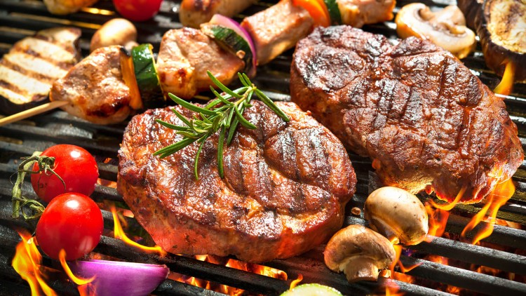 Verschiedene gegrillte Speisen, darunter Fleisch, Würstchen, Gemüsespieße, Maiskolben, Champignons, Tomaten und Zwiebeln auf einem Grill.