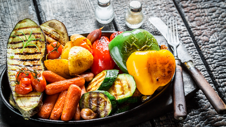 Gegrilltes Gemüse wie Aubergine, Paprika, Zucchini, Karotten, Kartoffeln und Tomaten auf schwarzem Teller, dahinter Besteck und Salzstreuer.