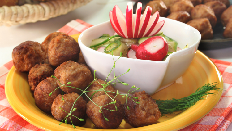 Gelbe Platte mit Fleischbällchen und Dill neben weißer Schale mit Gurken-Radieschen-Salat, dekoriert mit geschnitztem Radieschen.