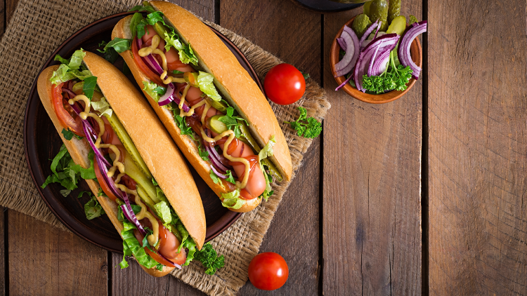 Zwei belegte Hotdog-Brötchen mit Salat, Tomaten, Gurken, Zwiebeln, Senf und Ketchup auf einem Teller, dekoriert mit Petersilie.