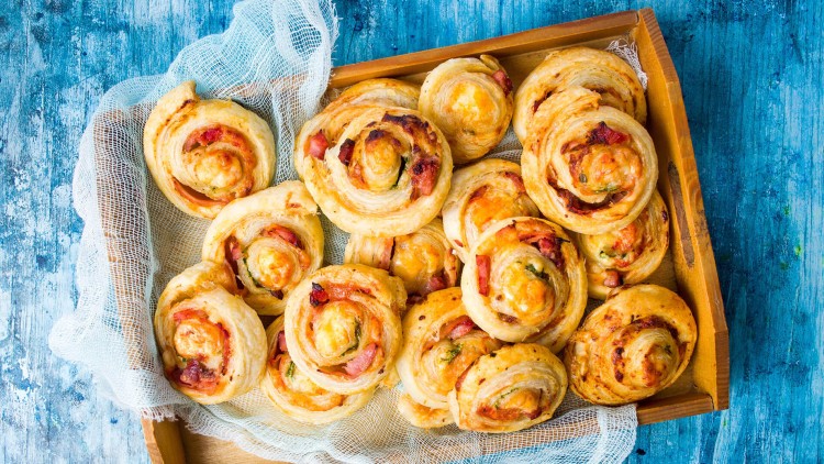 Mehrere goldbraune Blätterteig-Schnecken mit Schinken und Käse liegen auf einem hölzernen Tablett mit weißem Stoffeinsatz.
