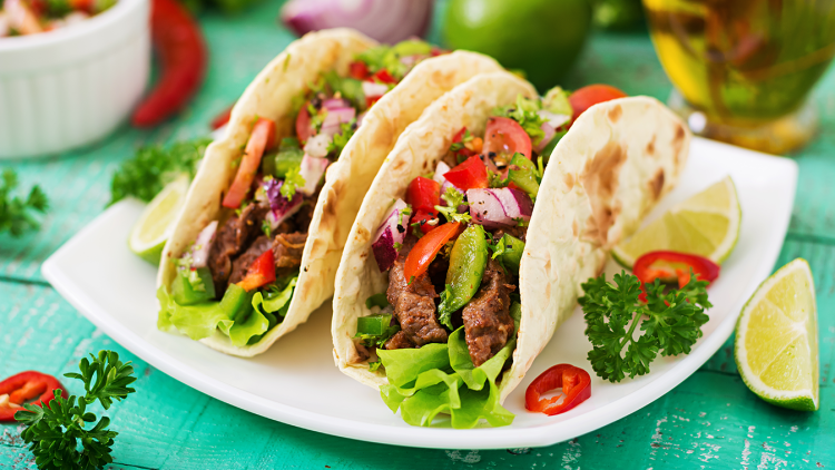 Drei gefüllte Weizentortillas mit Salat, Rindfleisch, Paprika, Tomaten, Zwiebeln und Limettenscheiben auf einem Teller.