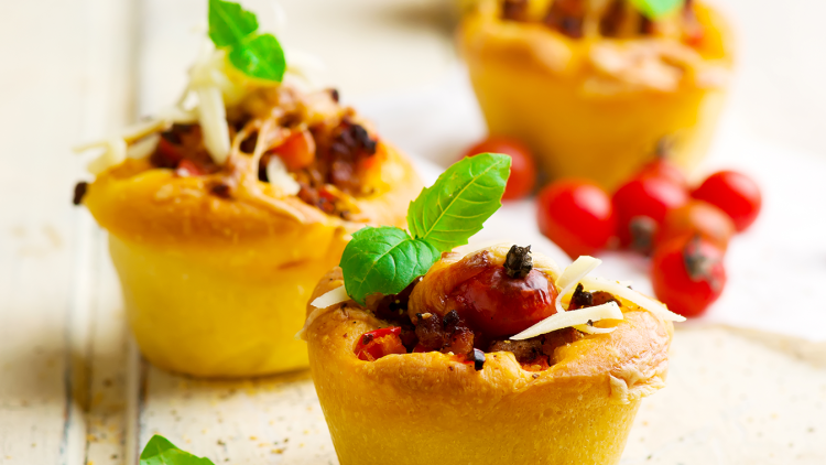 Herzhafte Muffins mit geschmolzenem Käse, Tomaten, Hackfleisch und Basilikumblättern auf heller Holzoberfläche.