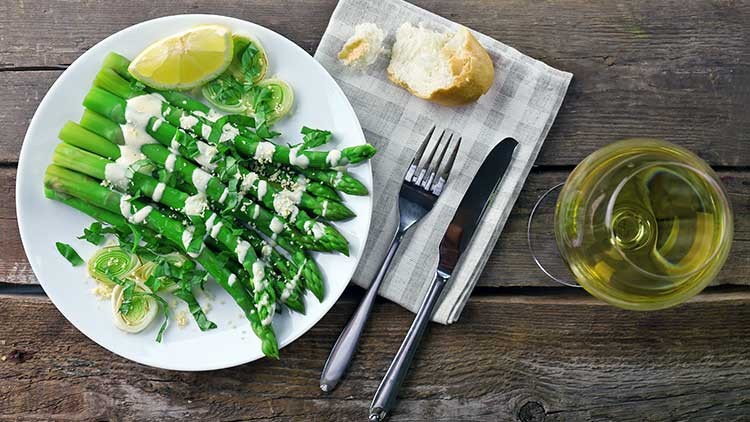 Ein Teller mit grünem Spargel, Zitronenscheibe und Sauce, daneben Besteck, Brot und ein Glas heller Wein auf Holztisch.