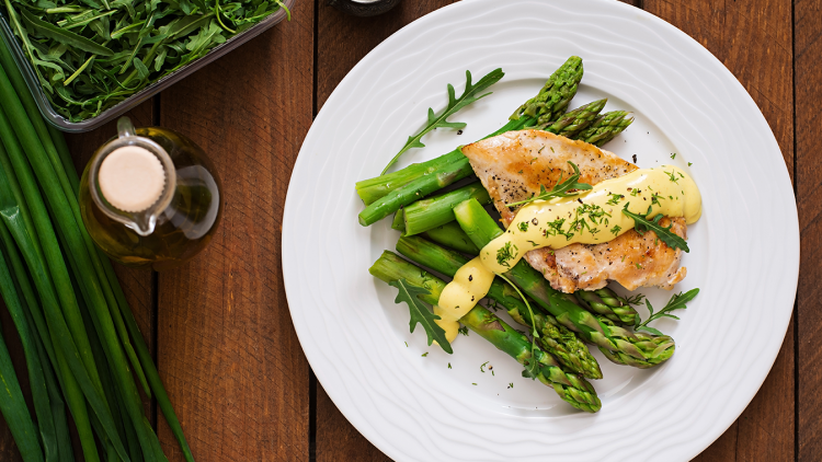Gegrilltes Hähnchen mit Spargel, Sauce und Kräutern auf einem weißen Teller, daneben Ruccola, Lauchzwiebeln und eine Ölflasche.