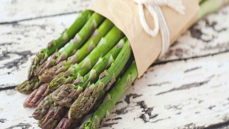Ein Bund grüner Spargel liegt in braunem Papier eingewickelt auf einer weißen Holzoberfläche mit dunklen Abnutzungsspuren.