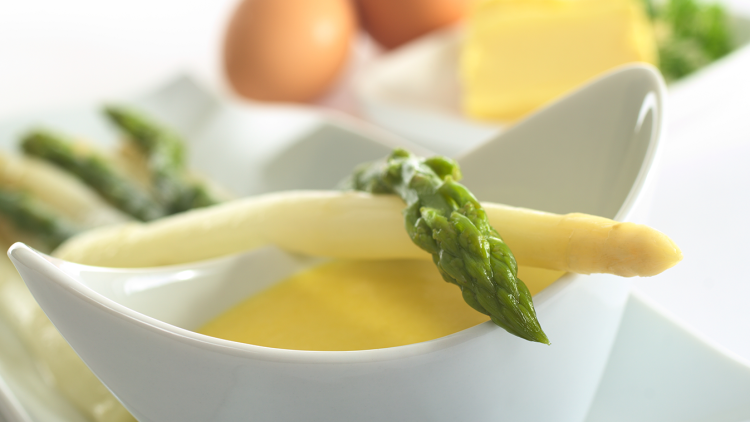 Weißer und grüner Spargel liegt auf einer weißen Sauce in einer Schale, im Hintergrund Eier, Butter und Petersilie.