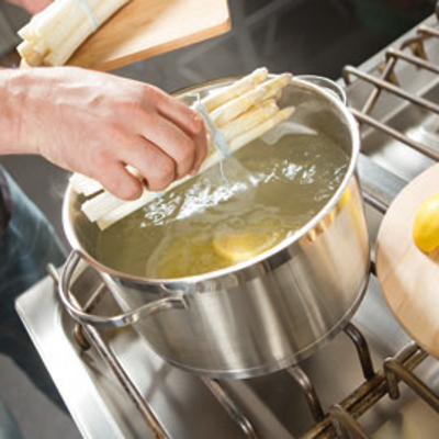 Eine Hand hält ein Bündel weißen Spargel über einen Topf mit kochendem Wasser, daneben liegt eine halbe Zitrone auf einem Brett.