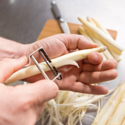 Eine Person schält weißen Spargel mit einem Sparschäler vor einem Holzbrett und einem Messer auf einer Metalloberfläche.