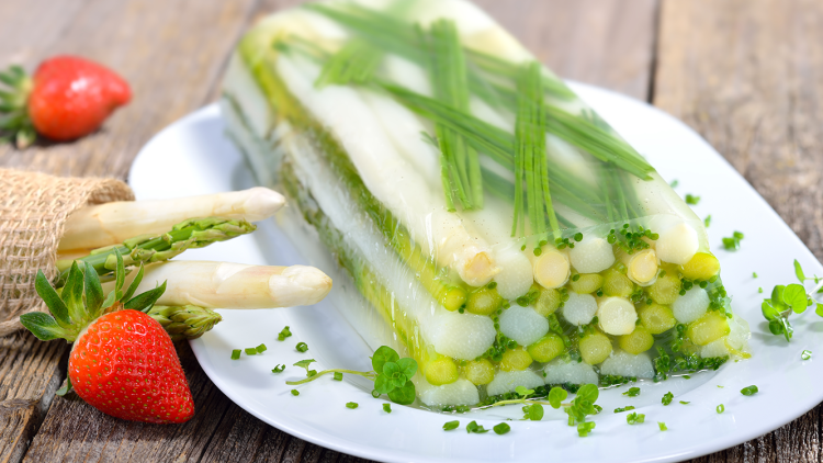 Ein rechteckiger Spargelterrine aus grünem und weißem Spargel mit Kräutern auf weißem Teller, daneben Erdbeere und Spargel.