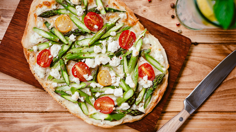Flammkuchen mit grünem Spargel, halbierten roten und gelben Kirschtomaten sowie weißem Käse auf Holzbrett serviert.