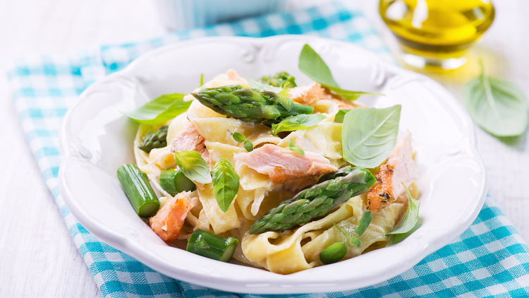 Bandnudeln mit grünem Spargel, Lachs und Basilikumblättern auf einem weißen Teller mit karierter Serviette.