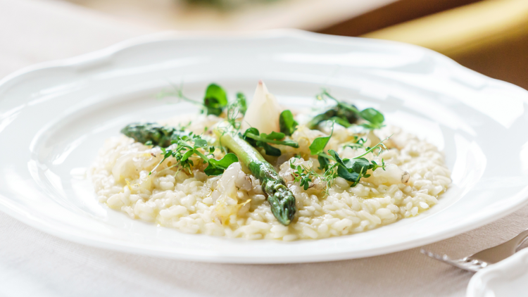 Ein Teller mit cremigem Risotto, garniert mit grünem Spargel, frischen Kräutern und kleinen weißen Zwiebelstückchen.