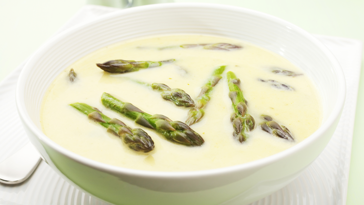 Weiße Keramiksuppenschale mit cremiger Suppe und grünen Spargelspitzen auf einem weißen Teller mit Löffel.