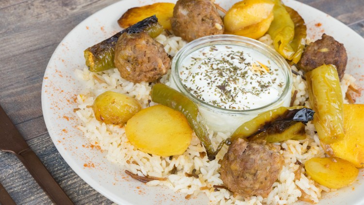 Ein Teller mit Reis, Fleischbällchen, gebratenen Kartoffeln, Grillpaprika und einer Glasschale mit Kräuter-Joghurt in der Mitte.