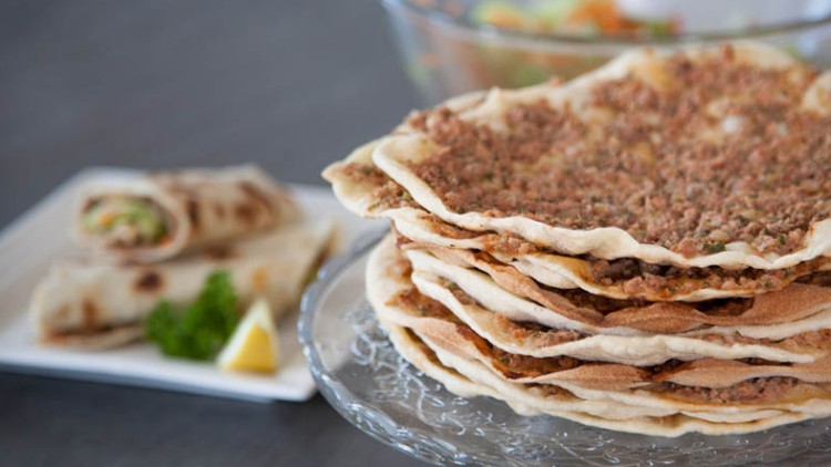 Ein Stapel dünner Fladenbrote mit Hackfleisch neben gefüllten Teigrollen, Petersilie und Zitronenspalte auf Tellern.