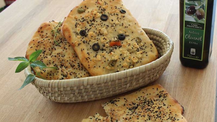 In einem Korb liegen belegte Fladenbrote mit Oliven und Sesam, daneben ein angebrochenes Fladenbrot und eine Flasche Olivenöl.