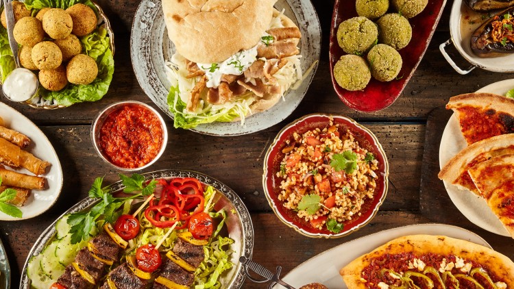 Vielfältige orientalische Speisen wie Falafel, Kebabspieße, Pide, Salate und Dips auf einem rustikalen Holztisch angerichtet.