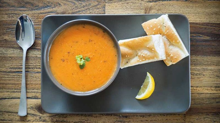 Graue Schale mit orangefarbener Suppe, dekoriert mit grüner Kräutergarnitur, Fladenbrot und Zitronenspalte auf einem Tablett.
