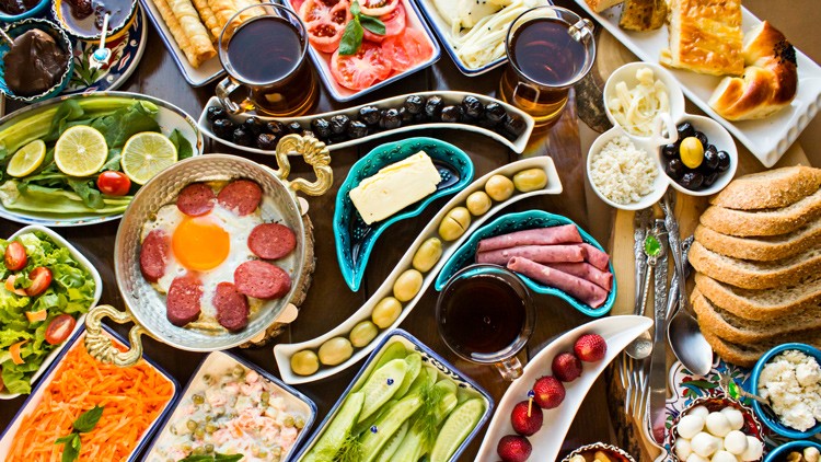 Frühstückstisch mit diversen Speisen wie Ei mit Wurst, Gemüse, Brot, Käse, Butter, Oliven, Tee und Fruchtdekor.