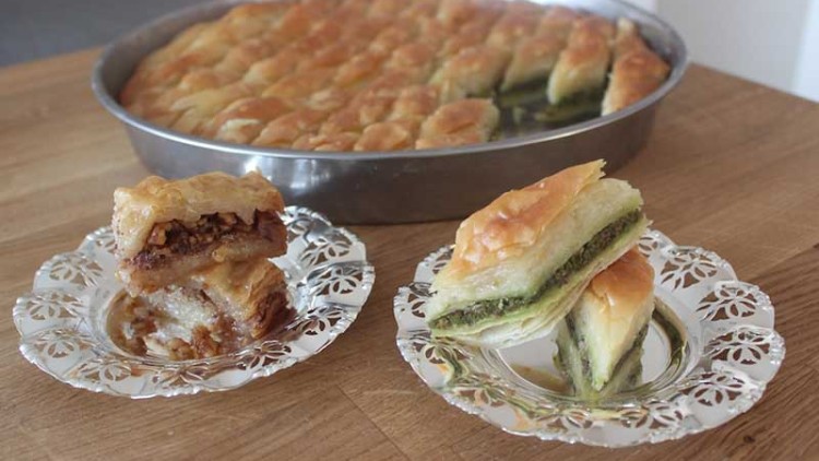 Eine runde Metallform mit Baklava, daneben zwei kleine Teller mit Baklava-Stücken in Goldbraun und Grünfüllung auf Holztisch.