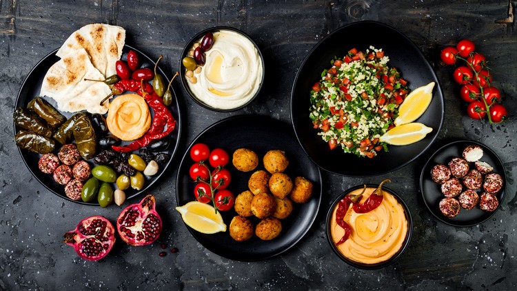 Mehrere schwarze Teller mit Falafel, Hummus, Taboulé, Dolma, Brot, Oliven, Granatapfel, Zitrone und Kirschparadeisern.