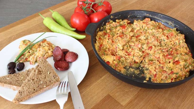 Omelette mit Tomaten und Paprika in einer Pfanne, dazu ein Teller mit Brot, Oliven, Wurst und frischen Tomaten auf Holztisch.