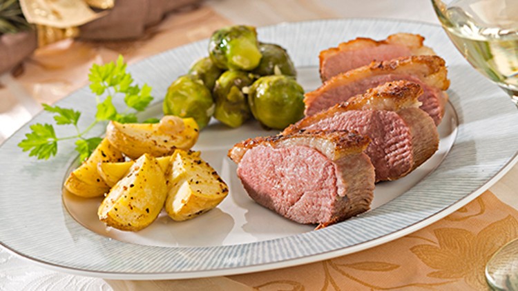 Ein Teller mit gebratenen Entenbrustscheiben, Kartoffelspalten, Rosenkohl und einem Zweig Petersilie auf weißer Tischdecke.