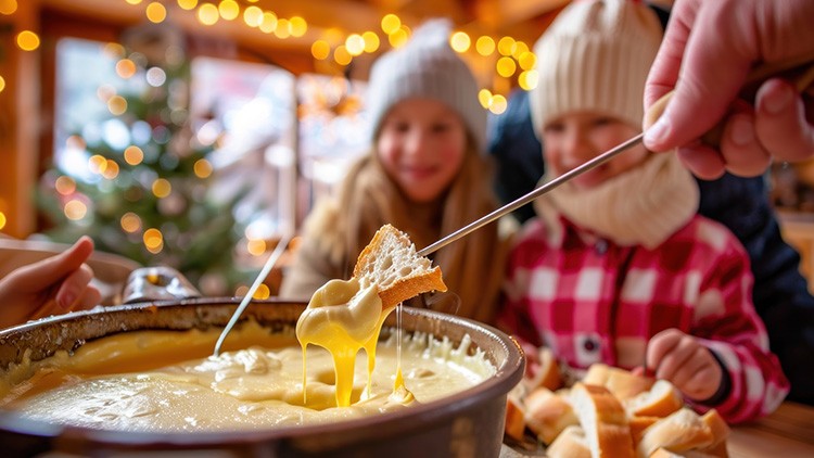 Ein Käsefondue in einem Topf, Brotstück wird mit einer Gabel eingetaucht, im Hintergrund Weihnachtsbeleuchtung und Baum.