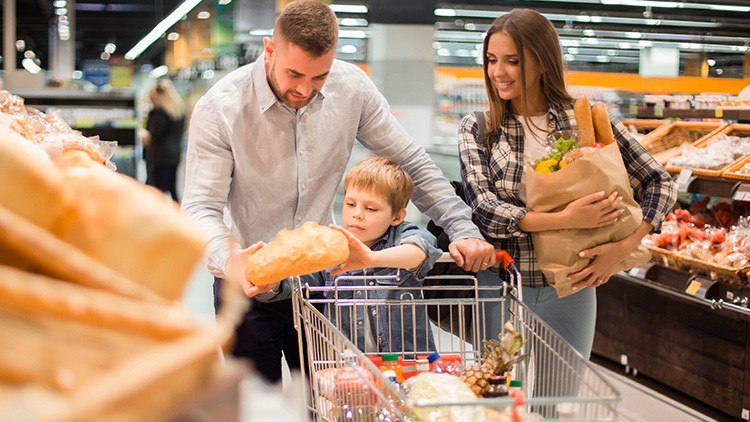Eine Familie steht an einem Einkaufswagen, das Kind greift nach einem Baguette, die Mutter hält eine Papiertüte mit Lebensmitteln.