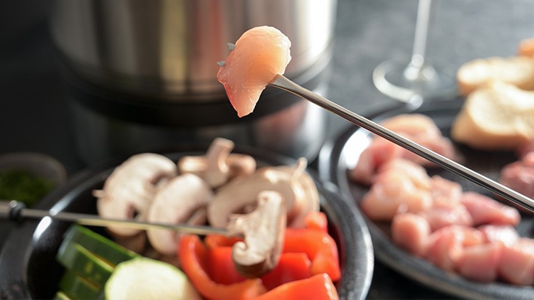 Ein Metallspieß mit Hühnerfleischstück, im Hintergrund Teller mit Gemüse, weiteren Spießen und ein Kochtopf.