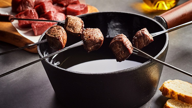 Vier Stücke Fleisch auf Metallspießen hängen über einem schwarzen Fondue-Topf, daneben liegen Brot und rohes Fleisch.