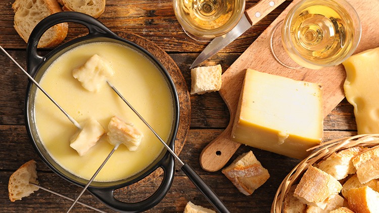 Fondue-Topf mit Gabeln, Käseblock, Brotwürfeln in Korb, Messer, zwei Weingläser und Brot auf Holztisch.