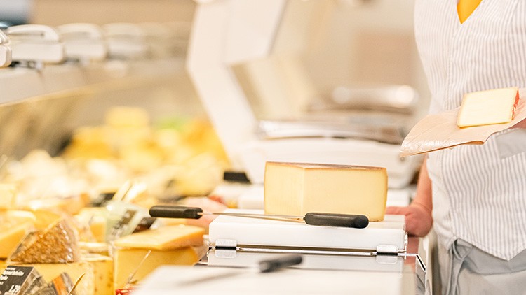 Käsetheke mit verschiedenen Käsesorten, ein Mitarbeitender hält ein Stück Käse auf einem Papier über einer Schneidemaschine.