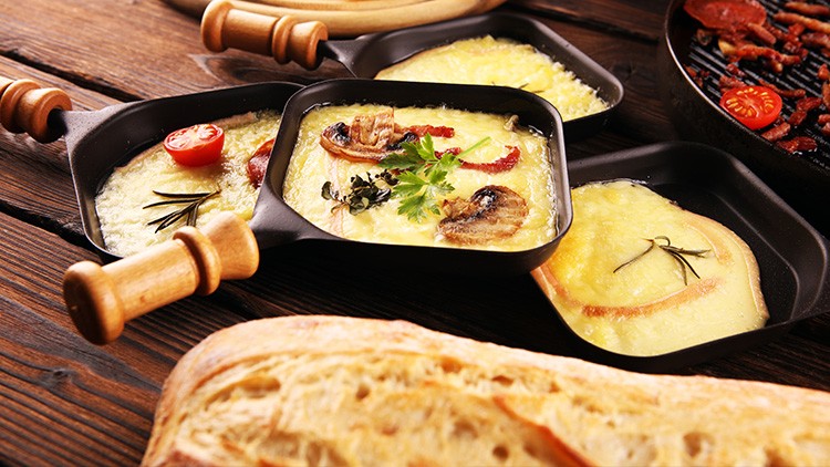 Drei kleine Pfannen mit geschmolzenem Käse, Pilzen, Tomaten und Kräutern, daneben frisches Brot auf Holzuntergrund.