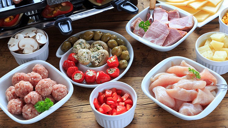 Verschiedene Zutaten für Raclette wie Käse, Schinken, Fleisch, Gemüse, Pilze, Oliven und gefüllte Paprika in Schalen.