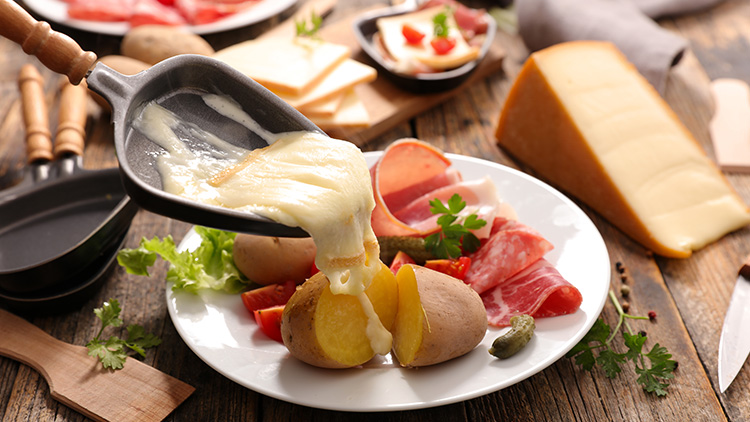 Schmelzender Käse läuft aus einer Pfanne über eine gekochte Kartoffel auf einem Teller mit Schinken, Tomaten und Beilagen.