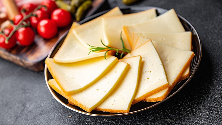 Auf einem Teller liegen mehrere Scheiben Käse, dekoriert mit einem Zweig Rosmarin, im Hintergrund Kirschtomaten und Gewürzgurken.