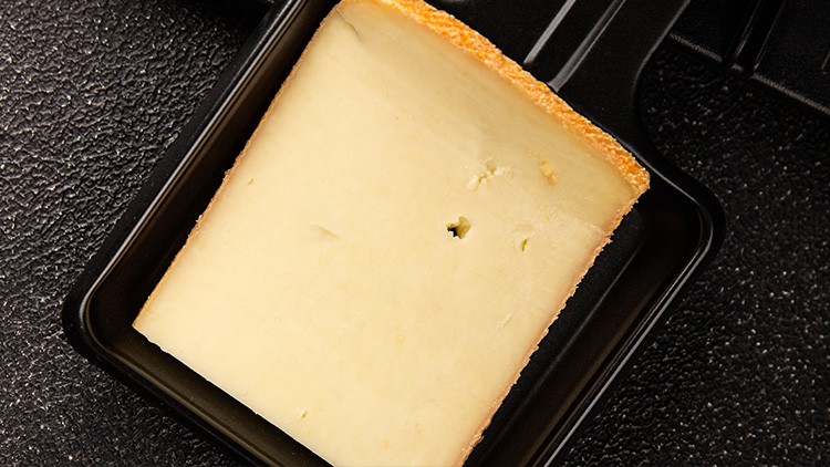 Ein rechteckiges Stück gelblicher Käse mit orangefarbener Rinde liegt auf einer schwarzen, glatten Unterlage.