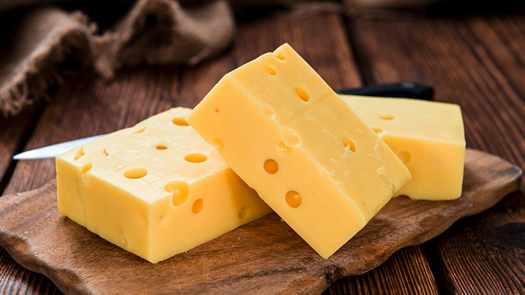 Drei Stücke gelber Käse mit Löchern liegen auf einem Holzbrett, im Hintergrund liegt ein Messer auf einer rustikalen Unterlage.