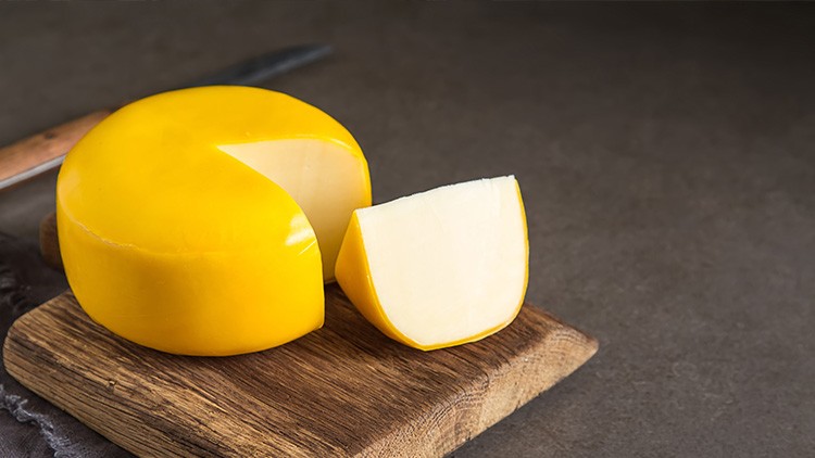 Gelber runder Käse mit glatter Oberfläche, angeschnitten auf einem Holzbrett, daneben ein Messer auf dunkler Oberfläche.