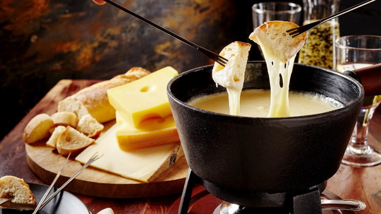 Brotscheiben auf Fonduegabeln werden in geschmolzenen Käse in einem schwarzen Fonduetopf getunkt, daneben Käse und Baguette.
