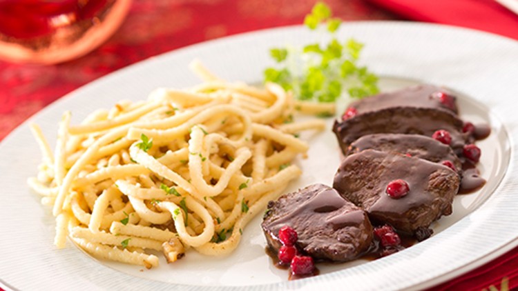 Portion Spätzle neben Scheiben von Fleisch in dunkler Sauce mit roten Beeren auf einem weißen Teller mit Kräuterdekoration.