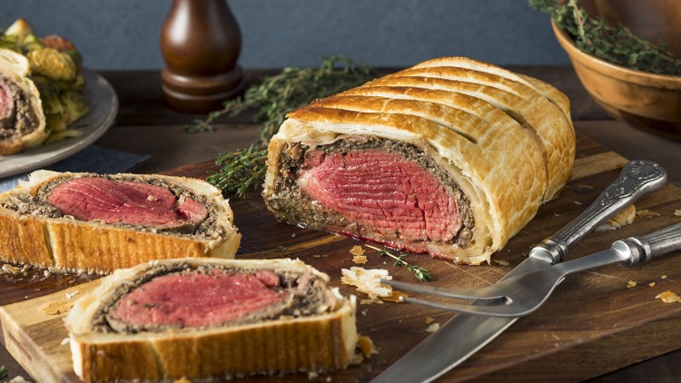 Anschnitt eines Beef Wellington mit rotem Kern auf Holzbrett, umgeben von Thymianzweigen, Messer und Gabel sowie einer Pfeffermühle.