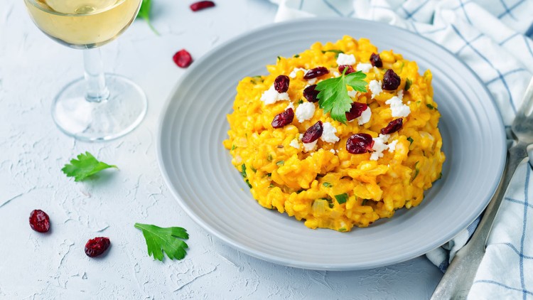 Ein grauer Teller mit gelbem Risotto, dekoriert mit Cranberrys, Petersilienblatt und weißem Käse, daneben ein Glas Weißwein.