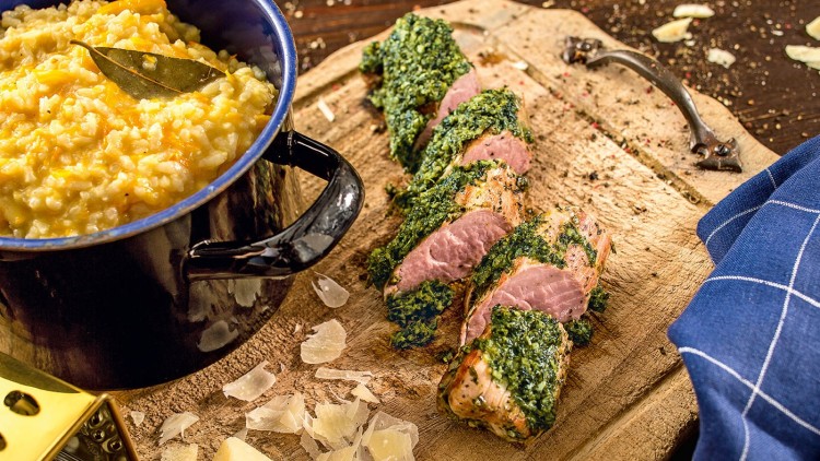 Ein Topf mit Kürbisrisotto und Lorbeerblatt steht neben Schweinefilet in Kräuterkruste auf einem Holzbrett mit Parmesan.
