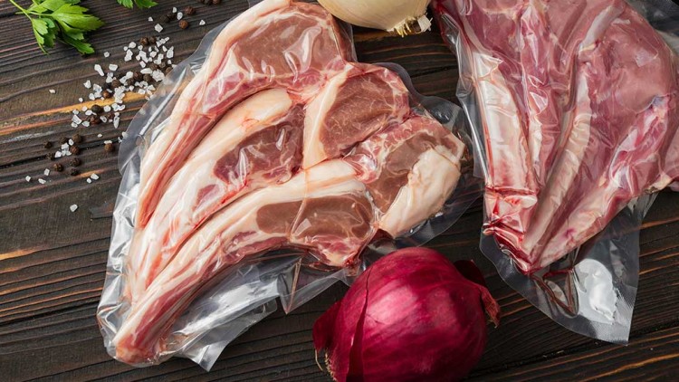 Zwei vakuumverpackte Lammkotelettstücke auf Holztisch mit weißen und roten Zwiebeln, Petersilie, Salz und Pfefferkörnern.